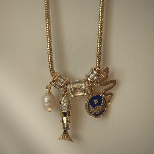 Gold charm necklace with a cobalt evil eye medallion, serpent, fish and freshwater pearl